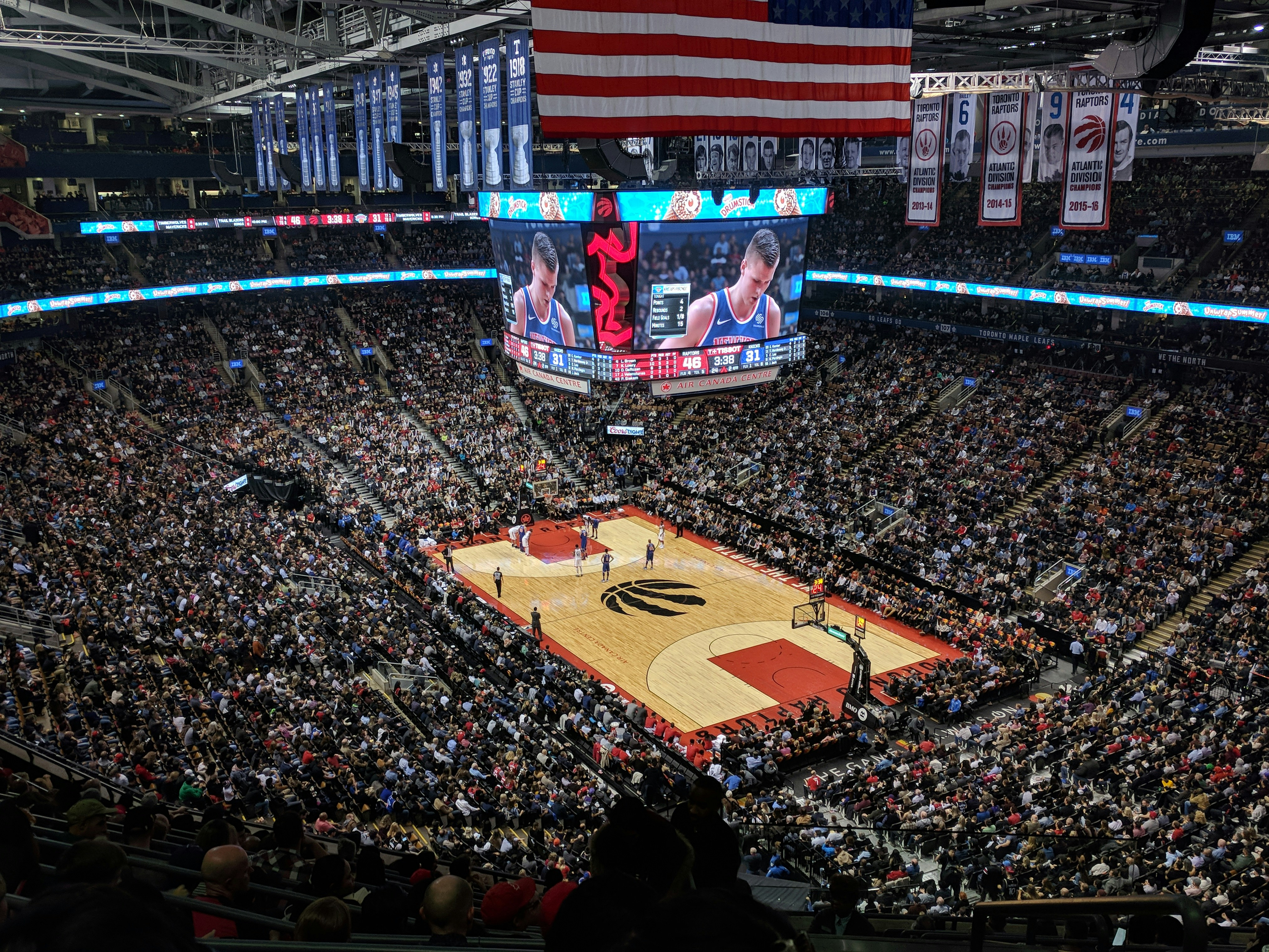 A basketball arena exterior at dusk with illuminated signage and a small crowd of fans walking toward the entrance.